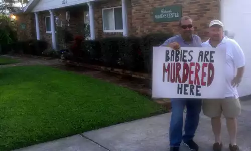 men standing outside planned parenthood with a sign that reads, "babies are murdered here."